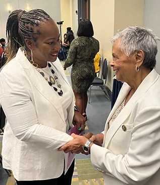 Two women in white jackets smiling and shaking hands at an indoor event (photo)