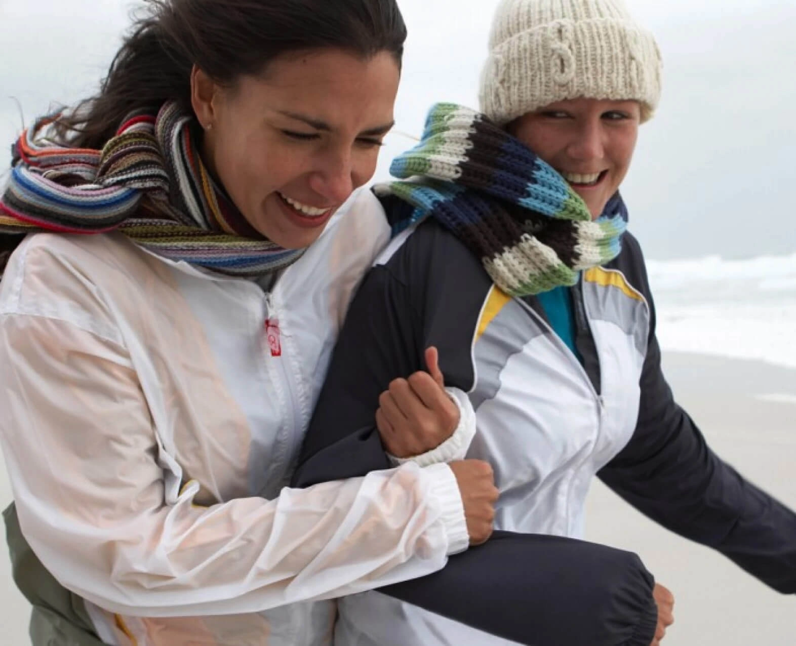 Two women bundled up in jackets and scarves, laughing and walking together on a windy beach. (photo)