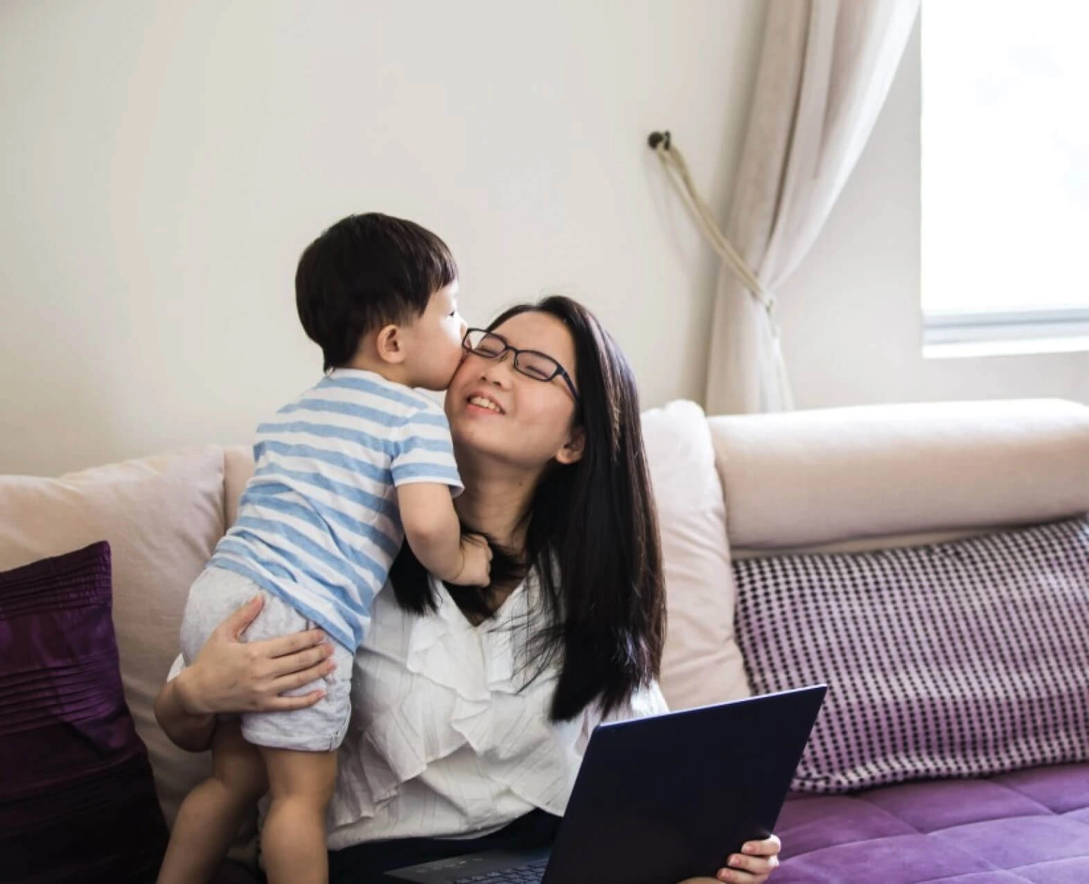 A young child giving a kiss to a woman holding a laptop, seated on a couch. (photo)